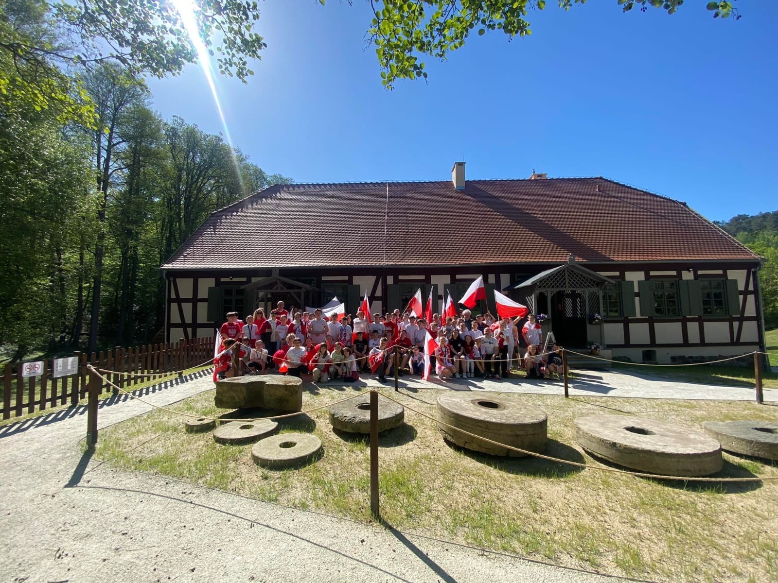 Uczniowie w biało czerwonych strojach z flagami polski pozują przed budynkiem młyna do zdjęcia.