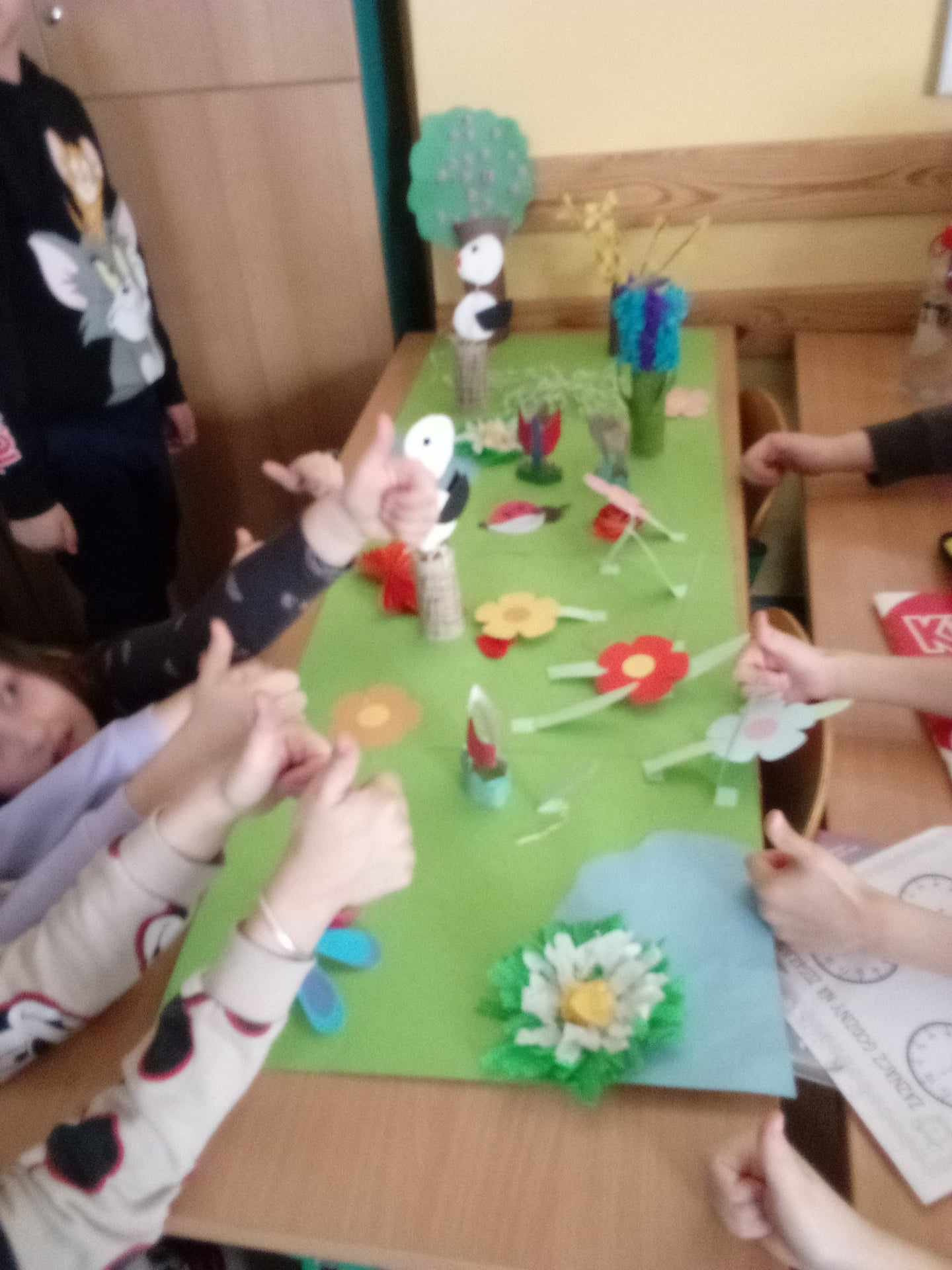 Przy ławce szkolnej na której rozłożone są przybory plastyczne siedzą uczniowie i wycinają kwiaty z kolorowego papieru. Nie widać ich twarzy ale ręce wskazują podniesiony kciuk co świadczy o zadowoleniu z tej aktywności.