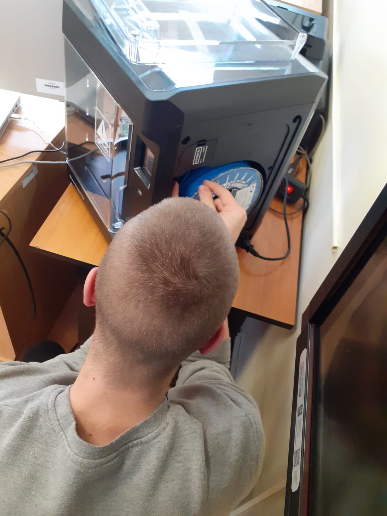 Kilkunastoletni chłopiec odwrócony tyłem  kuca przed drukarką 3D. Zakłada filament.