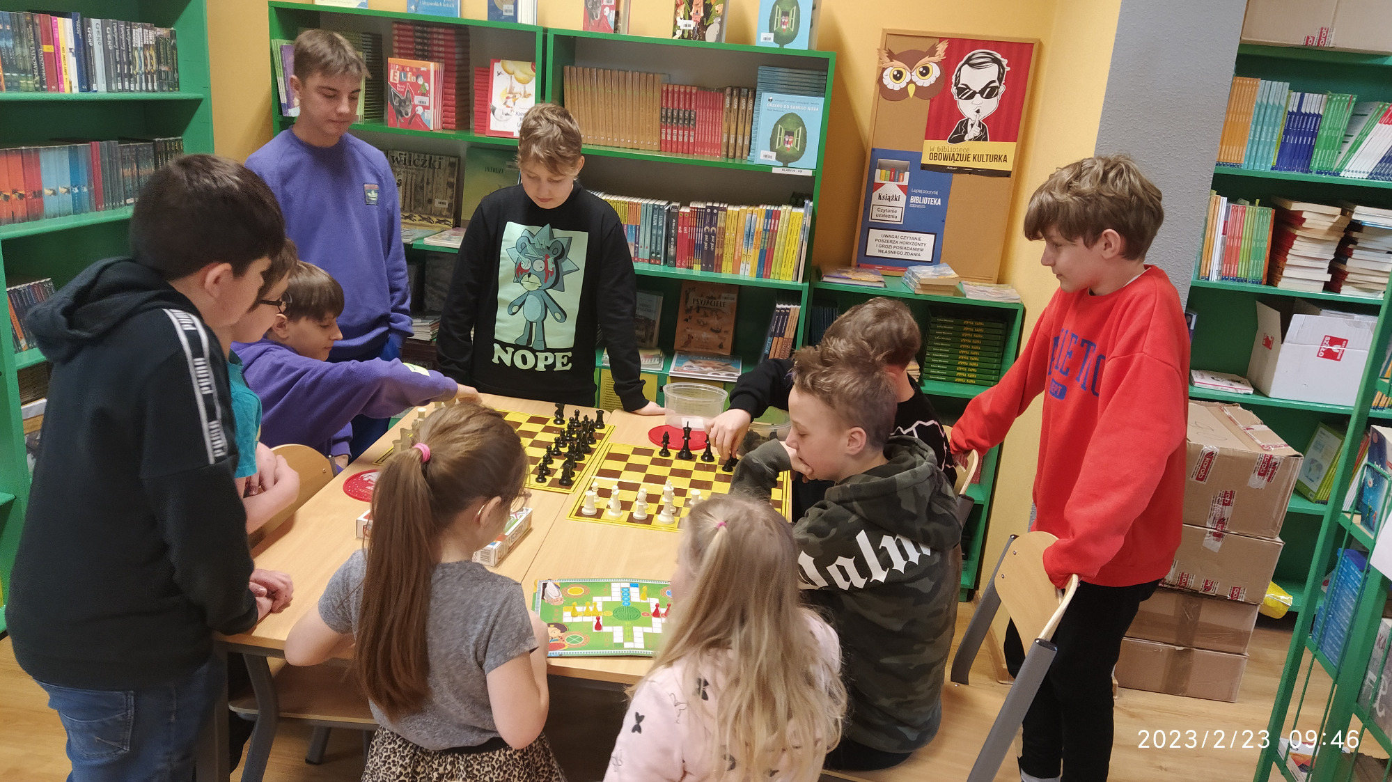 Uczniowie przy stoliku w bibliotece szkolnej grają w gry planszowe: szachy i Chińczyka.