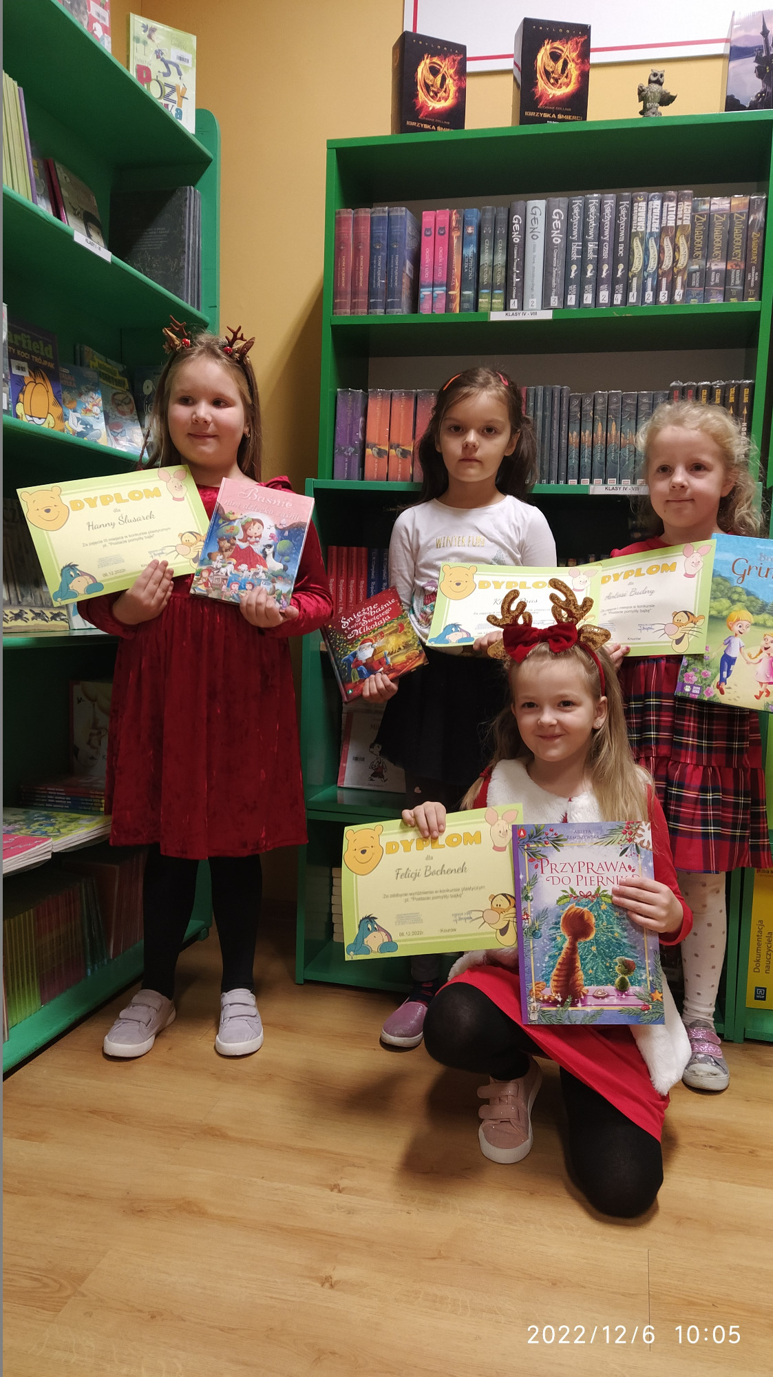 Laureaci konkursu czytelniczo-plastycznego w bibliotece  – cztery dziewczynki z dyplomami i nagrodami książkowymi. Za nimi regały z książkami.