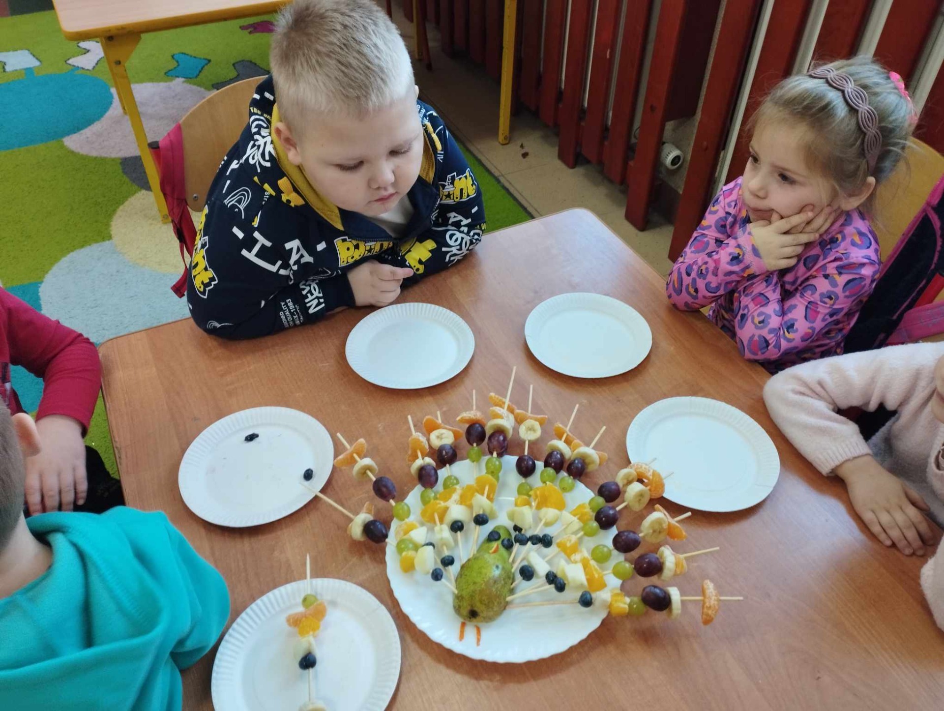 Przedszkolaki siedzą przy stoliku, na którym stoją talerzyki z owocowymi szaszłykami
