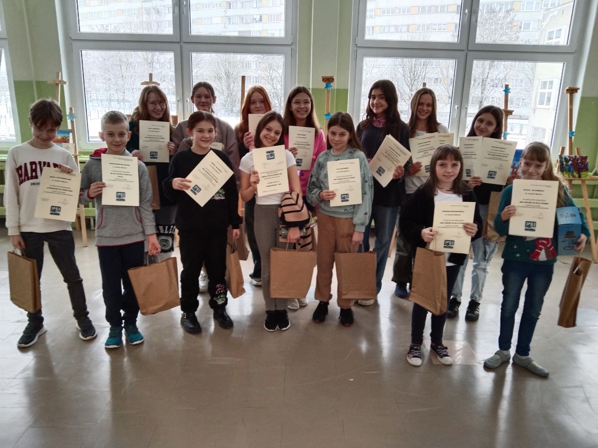 grupa uczniów stojąca na szkolnym korytarzu trzymająca szare torby  z nagrodami i dyplomy świadczące o sukcesach w konkursach
