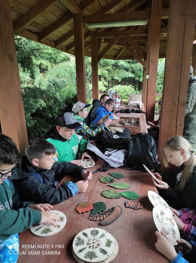 Uczniowie klasy czwartej siedzą pod wiatą przy stole i rozwiązują zadania ze znajomości tropów zwierząt, drzew oraz rozpoznania liści. W tle znajdują się drzewa.