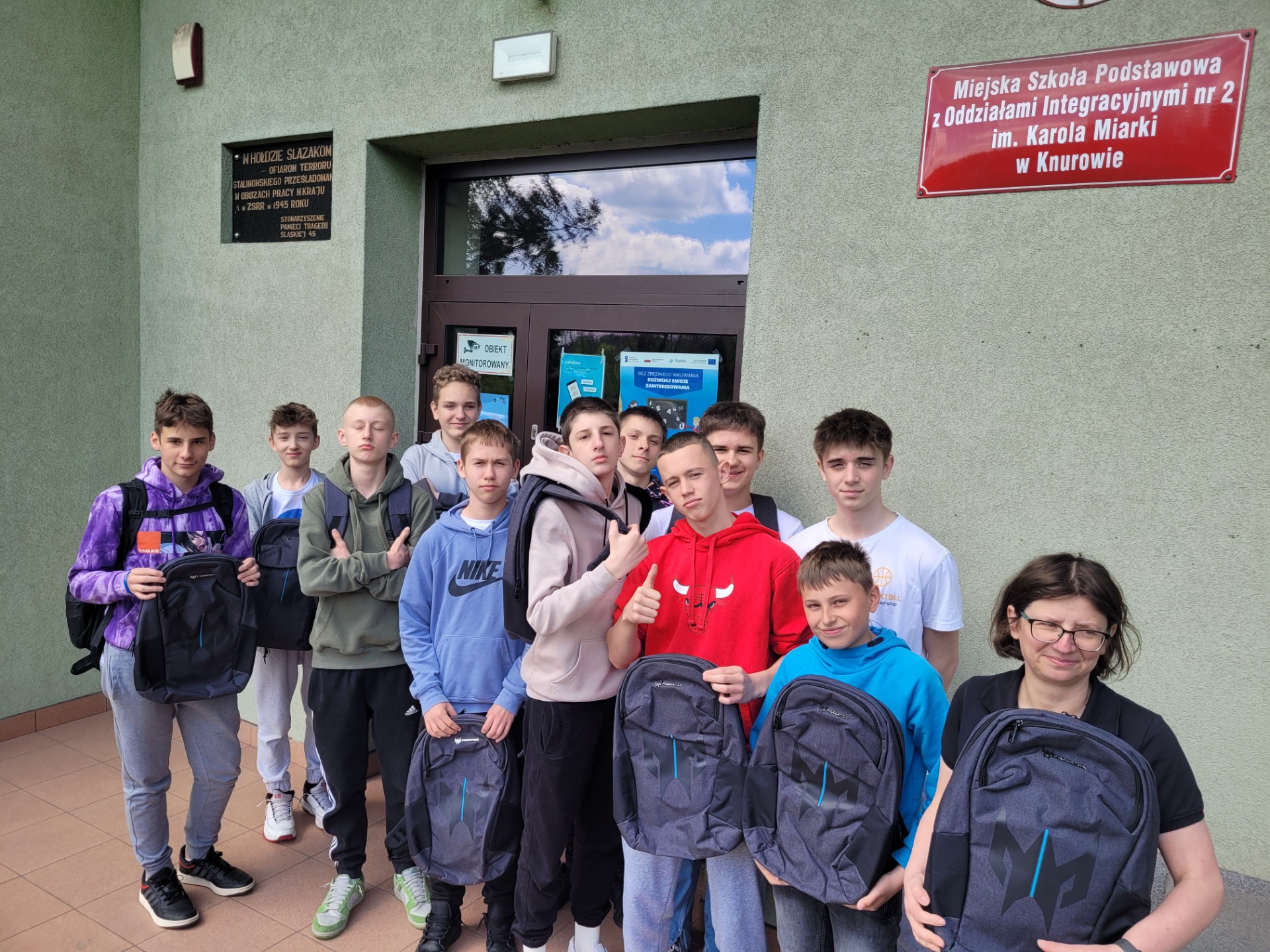 Grupa 10 uczniów wraz z nauczycielką trzymając plecaki uśmiechają się do zdjęcia przed budynkiem szkoły.