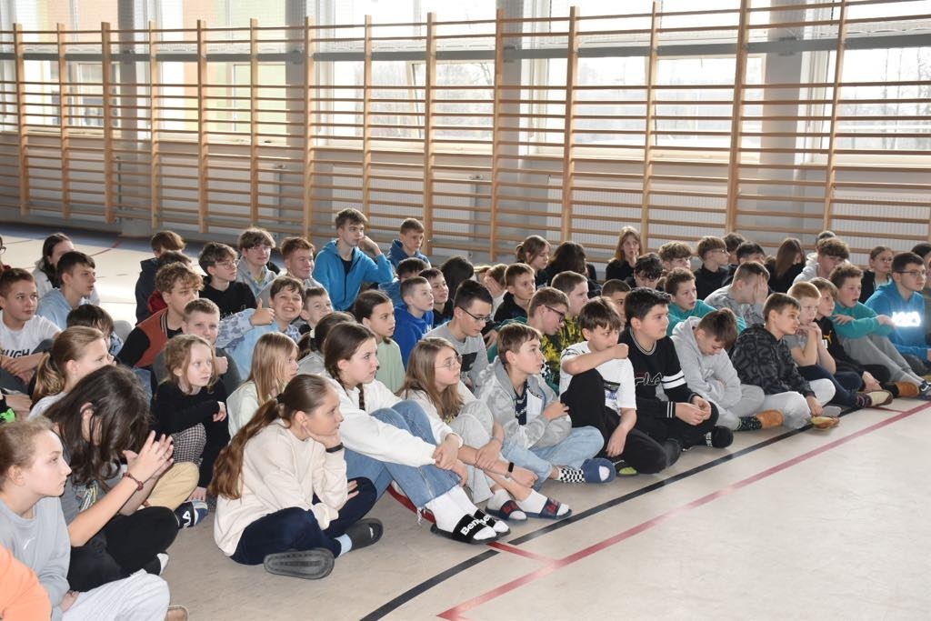 Grupa uczniów siedzi na podłodze w sali gimnastycznej.