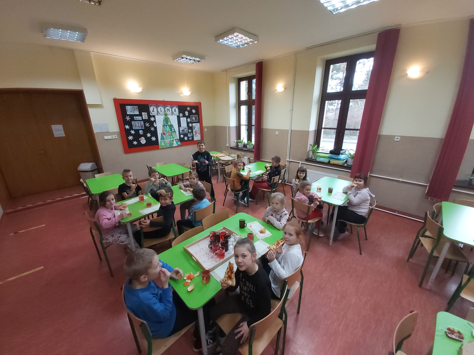 Dzieci siedzą przy stolikach w stołówce szkolnej i jedzą pizzę
