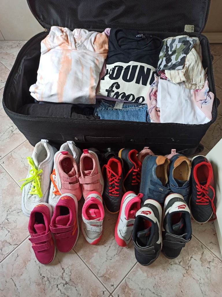 Zdjęcie przedstawia dużą, otwartą walizkę, w której znajdują się kolorowe ubrania. Przed walizką, na podłodze ustawiono parami kolorowe, sportowe obuwie różnych rozmiarów.