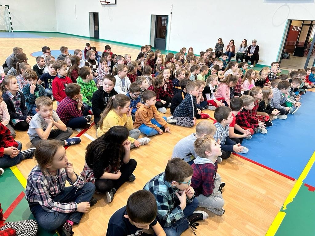 Uczniowie klas 0-III wraz z nauczycielkami siedzą na sali gimnastycznej. 