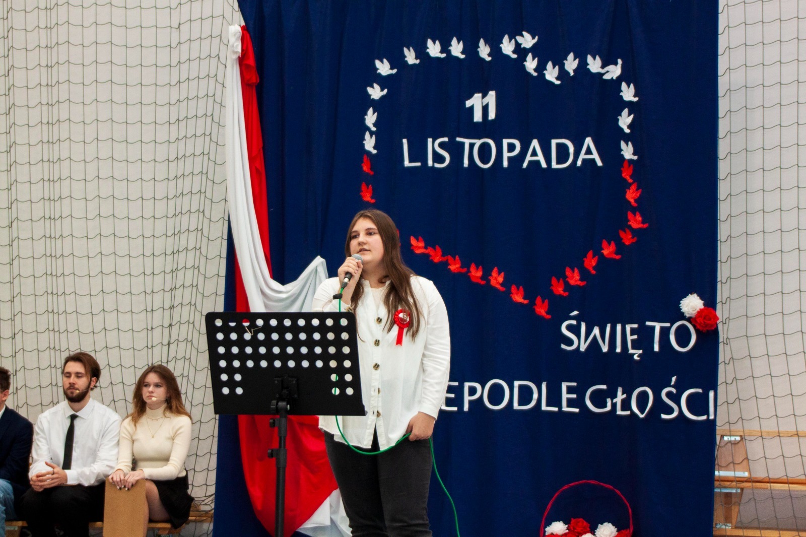 Uczennica w trakcie wystąpienia, w tle tablica z biało - czerwoną dekoracją. 