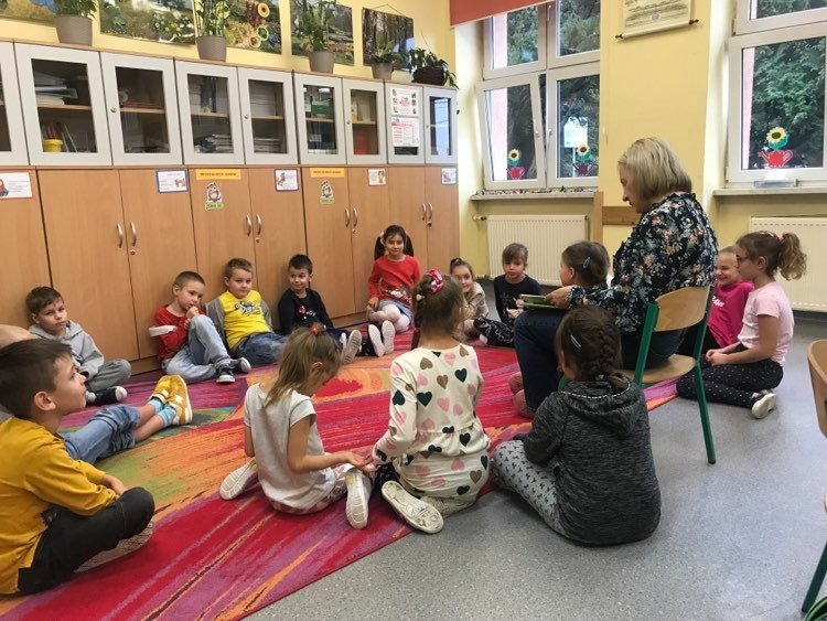 Dzieci siedzą na dywanie i słuchają czytanej przez nauczycielkę książki.